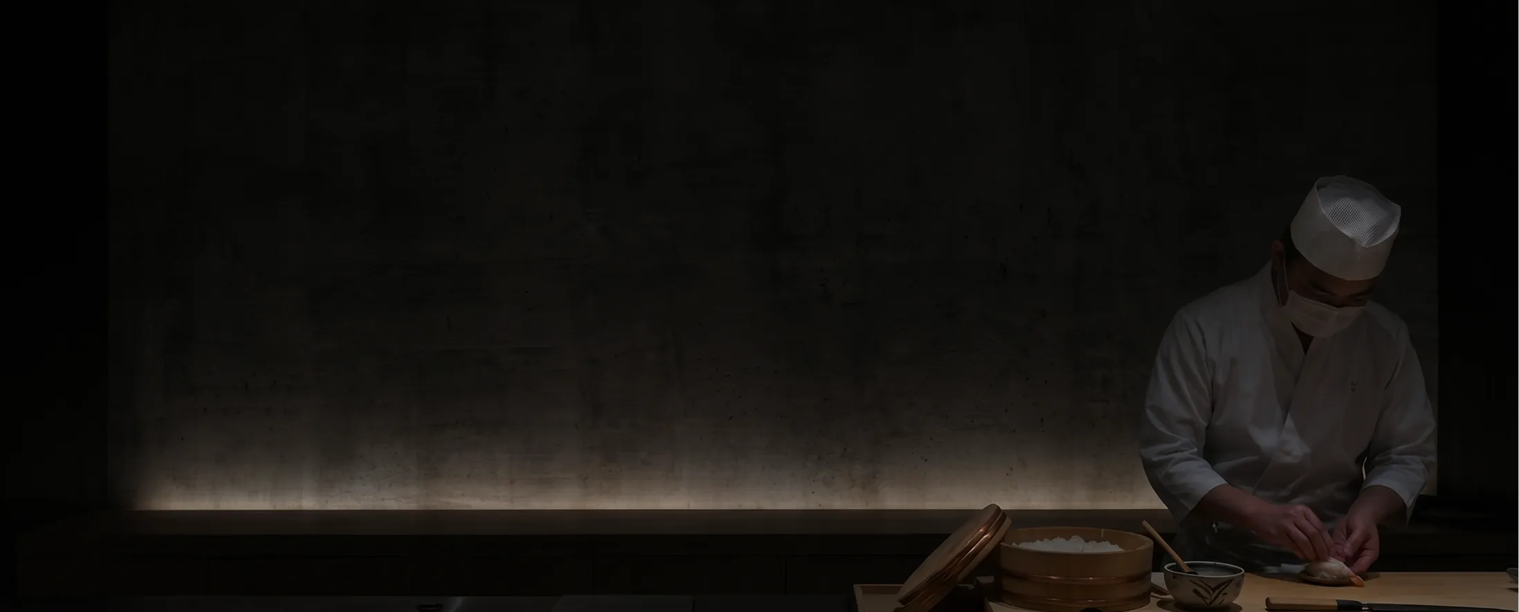 Sushi chef wearing white uniform and mask preparing sushi in a dimly lit restaurant.